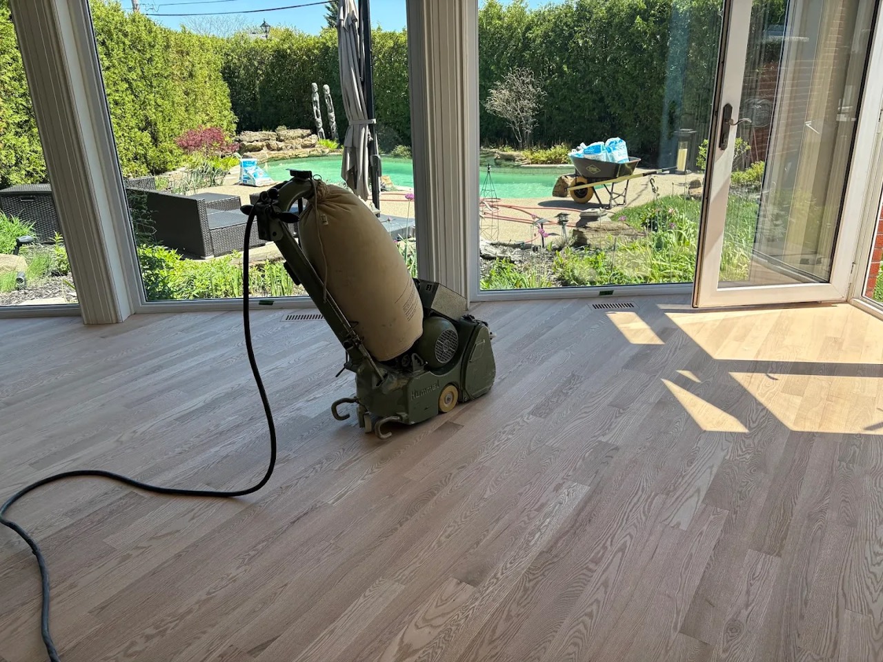 A Planchers Locas floor sander resurfacing a solid oak floor, with a bright backyard view